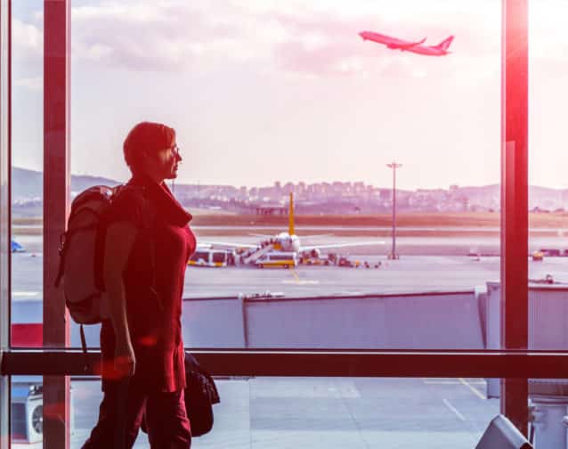 Woman with Hand Luggage Backpack in casual Clothing walking along large Window of Airport Terminal while Air Plane taking off behind