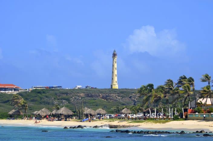 Arashi beach in Aruba with the California Lighthouse on a cliff in the distance, palm trees, sandy coast, blue water, and people enjoying the beach