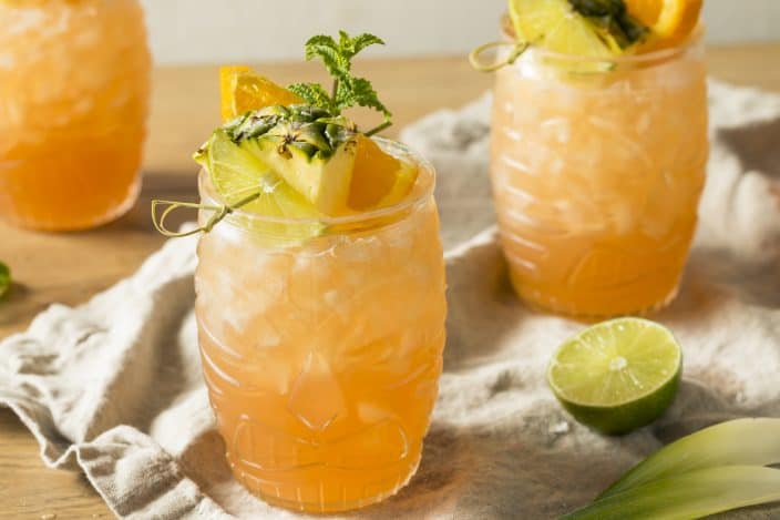 3 goombay smash cocktails in festive clear tiki classes with a garnish of pineapple, orange, and mint on top, sitting on a wooden table with a grey linen cloth on top and a half a lime lying next to them