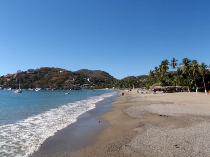 Beautiful tropical sandy beach with boats in the water and hills in the background in Zihautanejo, Mexico