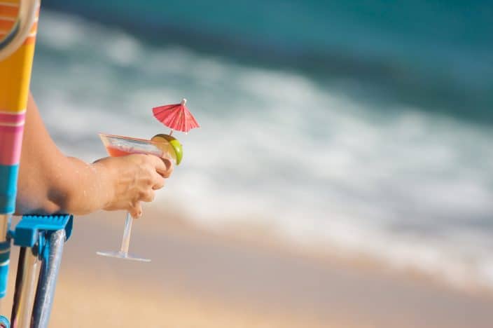 a woman holding a margarita glass with red drink umbrella and a lime sitting in a chair on the beach by the water during her beach day