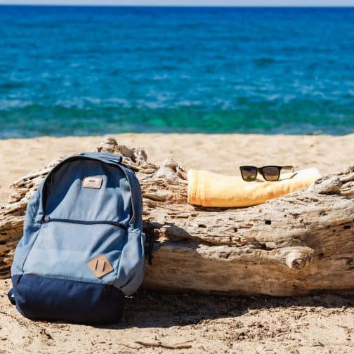 Picture of a blue backpack, towel, and sunglasses on a log with blue seashore and sand in the background. 