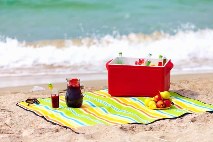 Picture of the beach with icebox placed on a multi-colored sheet with fruits on the side, chilled drink, sunglasses, and the waves all in the background. 
