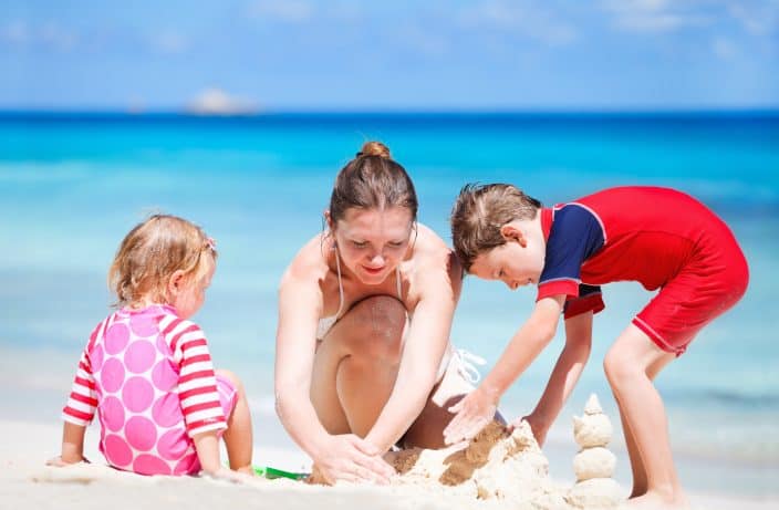 woman building sandcastle with her son in a red swimsuit and daughter in a pink swimsuit during her beach day with blue skies and blue water in the background. 