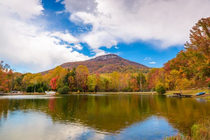  a lake in Georgia with trees on the banks with gold, orange, and red leaves on them, with a mountain in the background with red and orange foliage and bright blue skies with clouds, a fall bucket list destination.