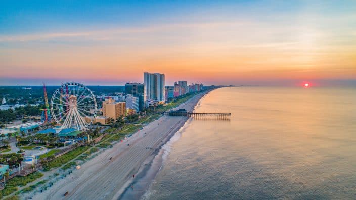 The shoreline in Myrtle Beach during sunset, with blue, pink and orange skies, a Ferris wheel with high rises on the beach and a pier going into the ocean. 