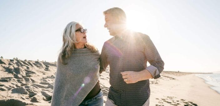 Woman with medium length hair and dark glasses, wearing a grey cover cashmere coverup with jeans while walking on the beach with her male partner wearing a checkered shirt and rolled sleeves making the perfect sunset evening attire to add to your packing list for beach vacation