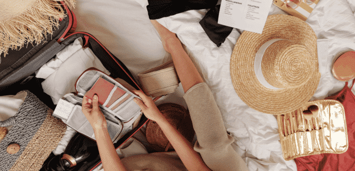 Packing suitcase with items for beach vacation. Wide brimmed hat on bed, passport and itinerary in her hands, makeup kit on bed beside her, packing list for beach vacation printed on bed beside her, suitcase open with clothing  placed inside and grey bag to carry items to the beach with. 