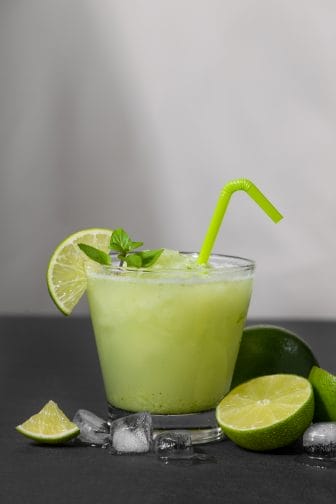 a green spicy frozen margarita with a green straw, limes, on a black background with limes scattered around the glass