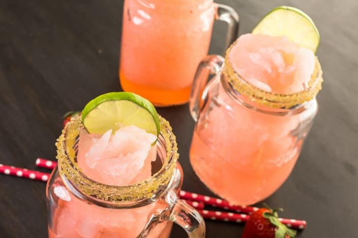 3 Frozen strawberry grapefruit margarita cocktails in drinking mason jars with lime slices and sugar on the rim on a dark wood table.