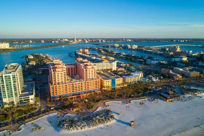 Aerial image of Clearwater Beach Florida, a romantic getaway weekend destination, with resorts and condominium apartments on the shoreline, bright blue skies and blue waters with bridges in the background. 