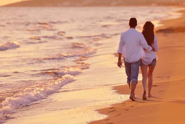 a man and a woman in white button down shirts and jeans walking on the beach during sunset to represent a romantic getaway weekend at the beach