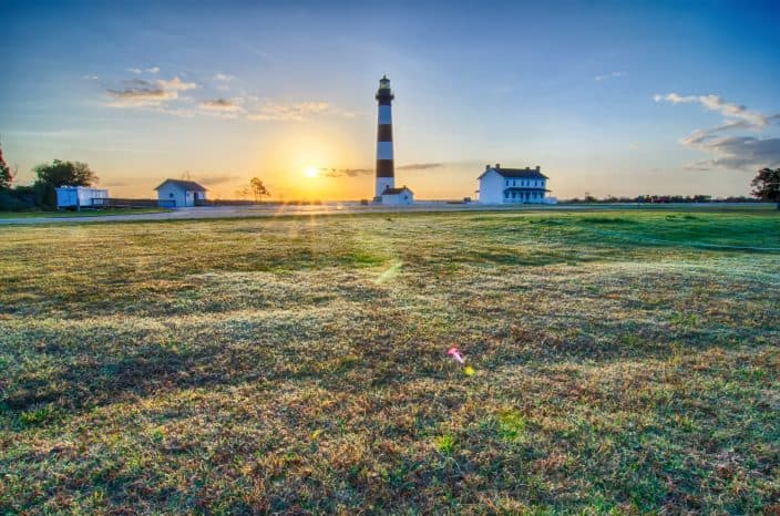  A large piece of farmland during sunrise with the Bodie Island Lighthouse and a white house next to it and a white shed on the left. The grass has dew on it and the sky is blue with an orange sun