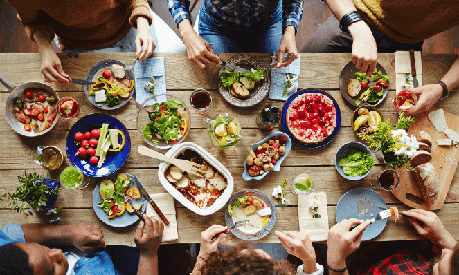 a overhead view of a bunch of people sitting at a wooden table with different plates of food and drinks on the table to represent summertime entertaining