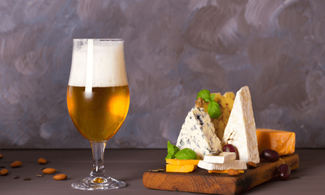 a large glass of beer next to a wooden board with different kinds of cheese wedges on it in front of a painted brown and blue background