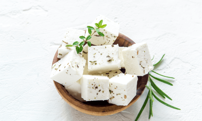 close up of feta cheese cubes in a wooden bowl with herbs on a white cement background