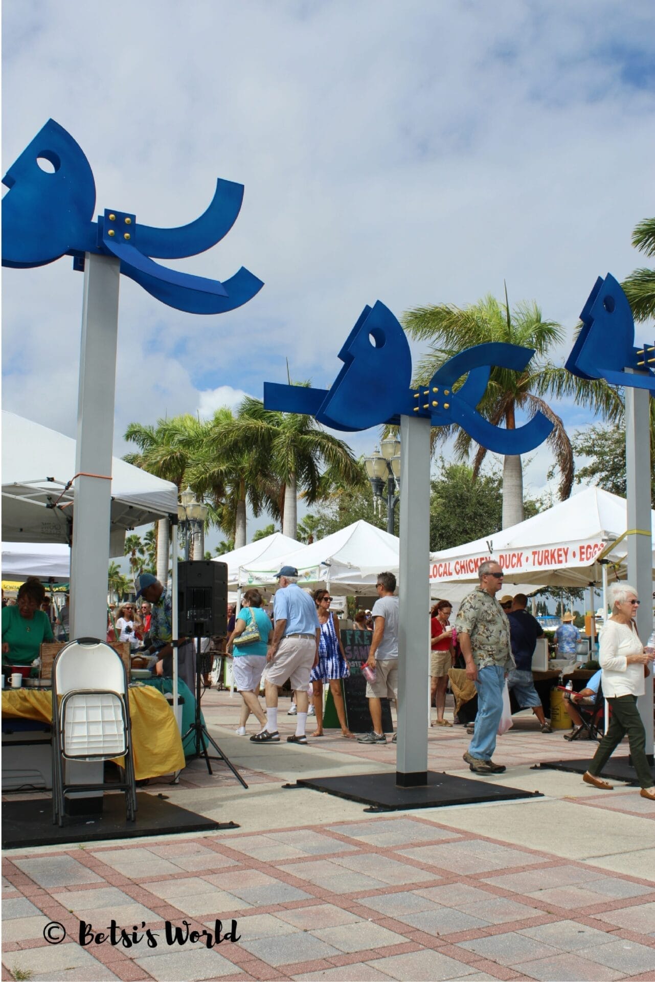 A farmers market in Fort Pierce with many people and tent stands and art sculptures.