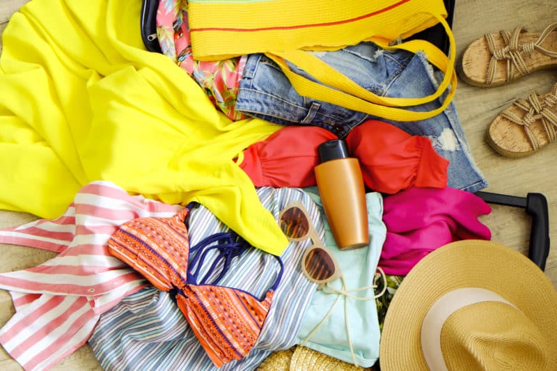 Open suitcase with pile of unfolded clothing on the floor. Woman packing for tropical vacation concept. Multiple unpacked female clothing items prepared for travel. Background, close up, copy space. What to pack for your romantic Bahamas getaway