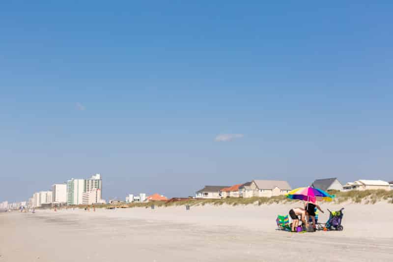 family on beach under striped umbrella, beach houses, blue skies and hotels on dunes in Myrtle Beach, a South Carolina romantic getaway