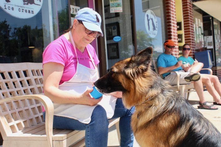 dog eating ice cream from cup in front of shop with woman in apron, pink shirt and jeans feeding the dog ice cream