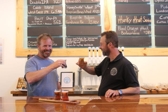 2 men toasting with beer behind counter with beer choices on chalkboard behind them and taps on the counter