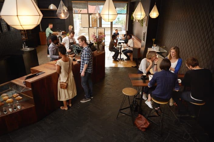High angle shot of a modern coffee shop in which there are baristas making coffees behind the counter, customers waiting at the counter, and other customers sitting at a table and talking. Visit local coffeeshops during your romantic staycation