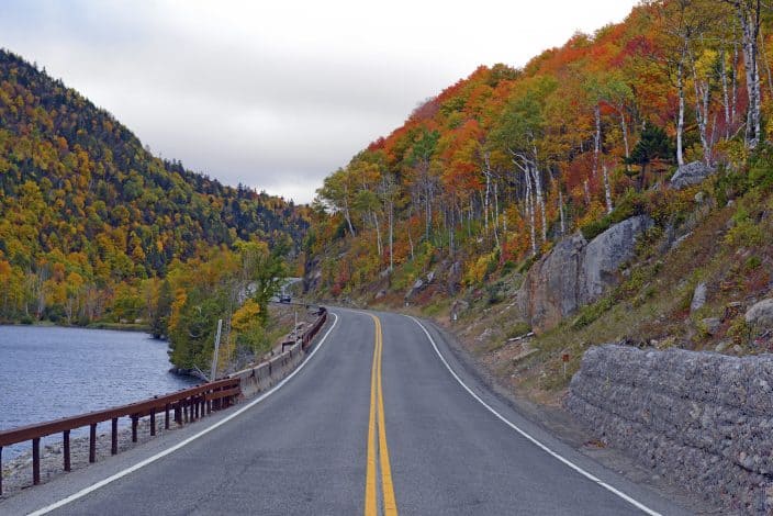 a road running through the mountains during fall, with trees with red, orange, and yellow leaves on the right side. There is a lake on the left with a guardrail against the road, as a representation of a fall road trip 