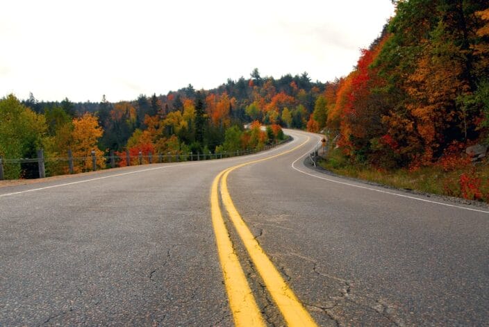 A winding road through the mountains, trees with fall foliage are on either side, an example of what you can see during a fall road trip