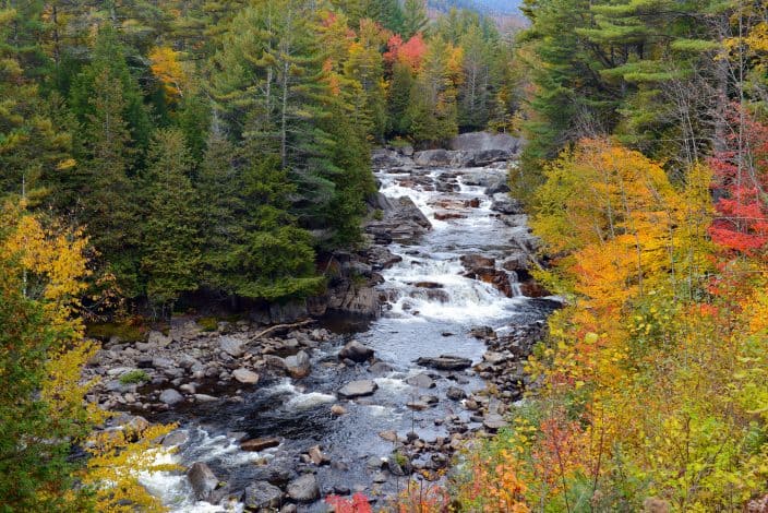 a creek filled with large and small grey rocks, running through the mountains with red, orange, yellow, and green foliage on the trees on either side, as a representation of what you can see during fall road trips