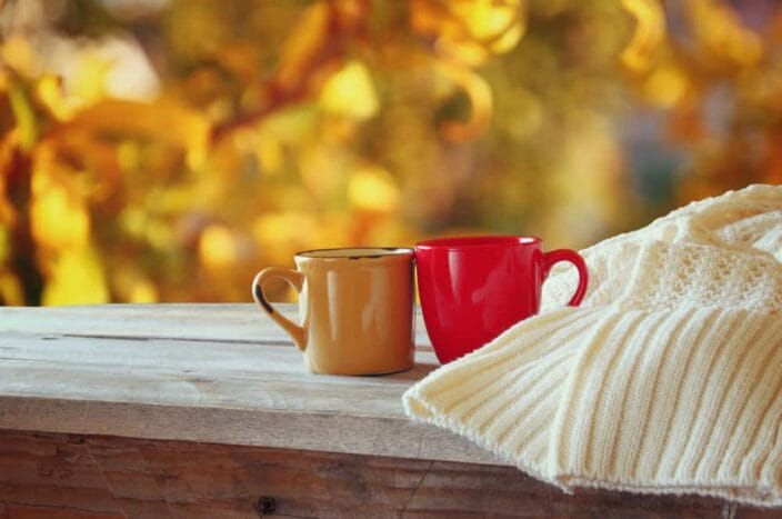 one orange mug and one red mug sitting next to each other on a wooden table with a cream knit sweater next them and fall foliage colors in the background, as a representation of a romantic getaway on a road trip