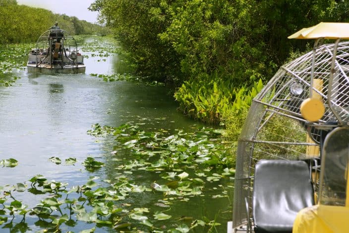 2 airboats in Everglades Florida Big Cypress National Preserve with lily pads on the water, green brush on the sides of the water and grey skies