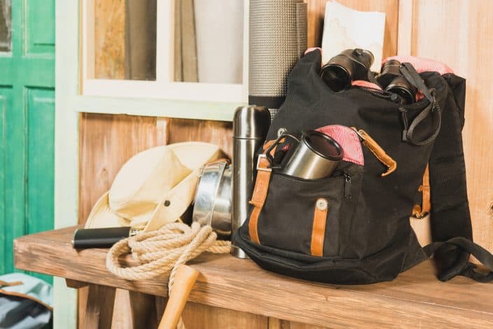 a black backpack sitting on a wooden bench with binoculars, a thermos, a rope, and a cowboy hat