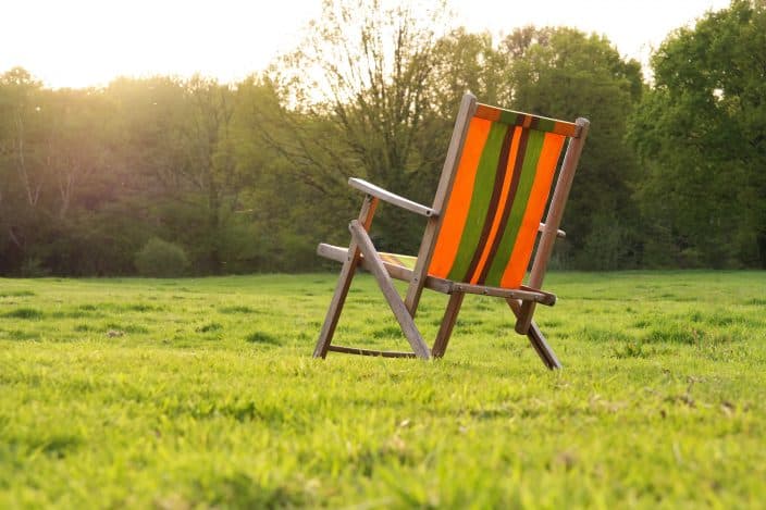 an orange and green striped chair with wooden legs sitting in a green field with trees during sunrise