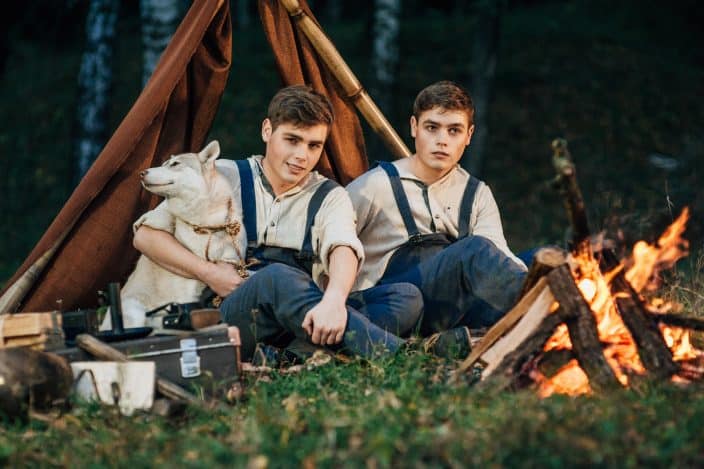 two twin brothers in blue jean overalls and beige shirts sitting in front of a campfire and in front of a brown tent with a dog at night