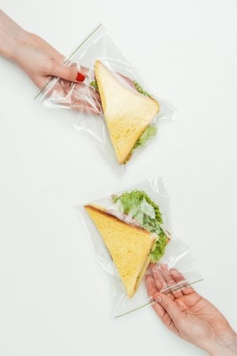 2 Ziploc bags with triangle cut sandwiches with lettuce on them inside of them on a white background. A hand with red nail polish is holding the top bag and another hand is holding the bottom bag.