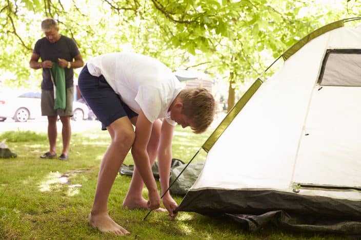 Father And Teenage Son Putting Up a white Tent, on green grass, with trees in the background On Camping Trip