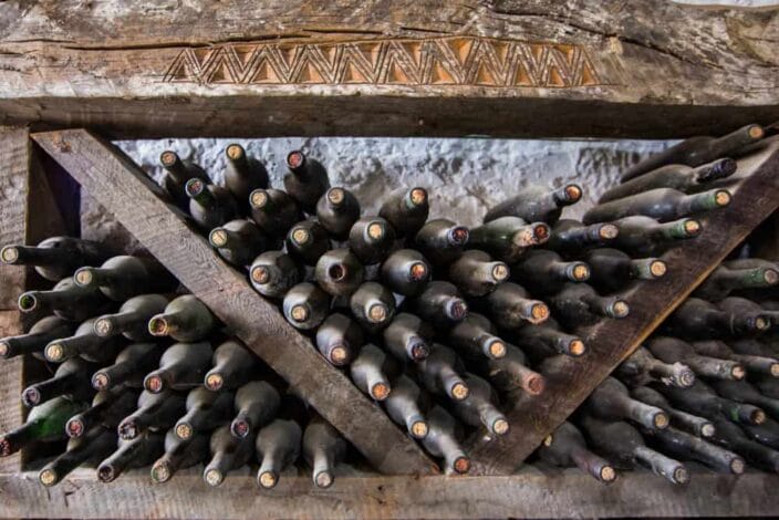 old, dusty wine bottles stack on top of one another in a old wooden shelf with a stone wall behind it  