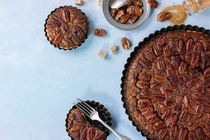 Homemade Big round maple pecan pie and small tartlets in black iron forms, served with brown sugar, maple and vintage cutlery over blue textured background.