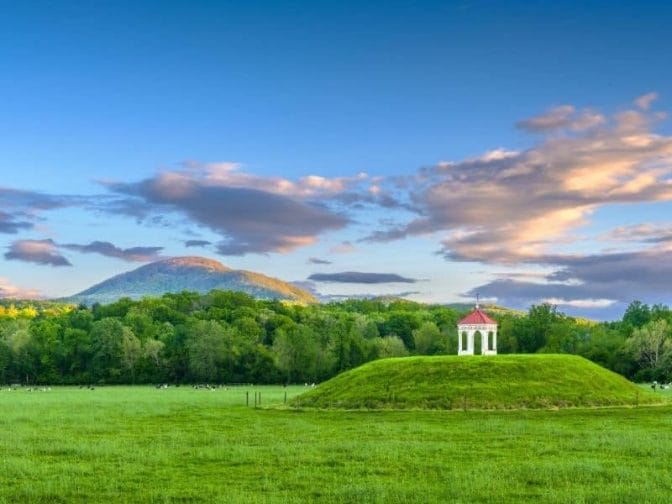 An Nacoochee Indian archeological site in Georgia, with blue skies, sunset clouds, a green field with a mound filled with grass and a white gazebo on top, an example of where to go during Georgia romantic getaways