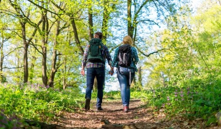 a man and a woman walking hand in hand up a mountain with green trees and blue skies to represent summer packing list for outdoor travel