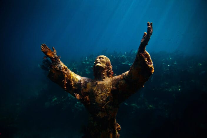Statue of Christ with his arms reaching up under the blue water with a background of blue water and green sea grass.