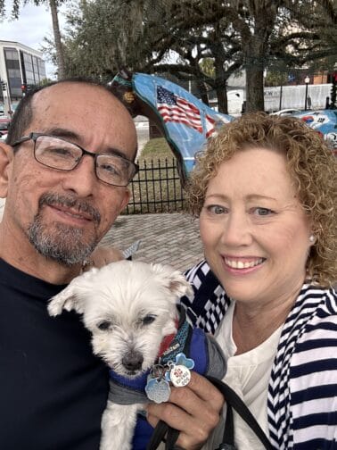 Betsi, of BetsiWorld & Betsi Hill Travel with Jim and with a white pup in front of a horse sculpture that is painted with American and Florida flags